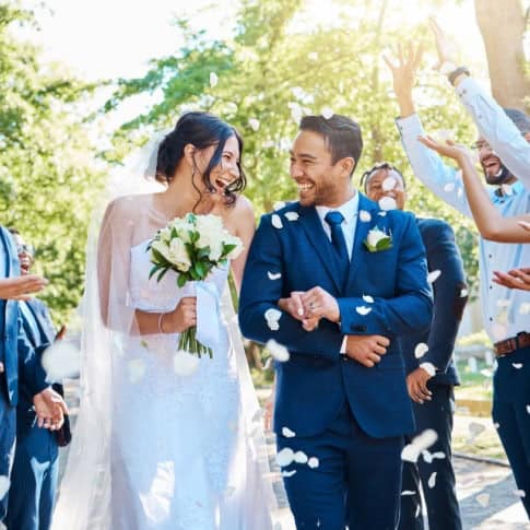 Bridal wedding celebration outdoors with smiling bride and groom, joyful guests, and flower petals.
